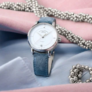 Armbanduhr Teutonia IV Lady mit pastellblauem Zifferblatt am blauen Veloursband steht auf hellblauem und rosafarbenem Stoff mit silbernem Schmuck.
