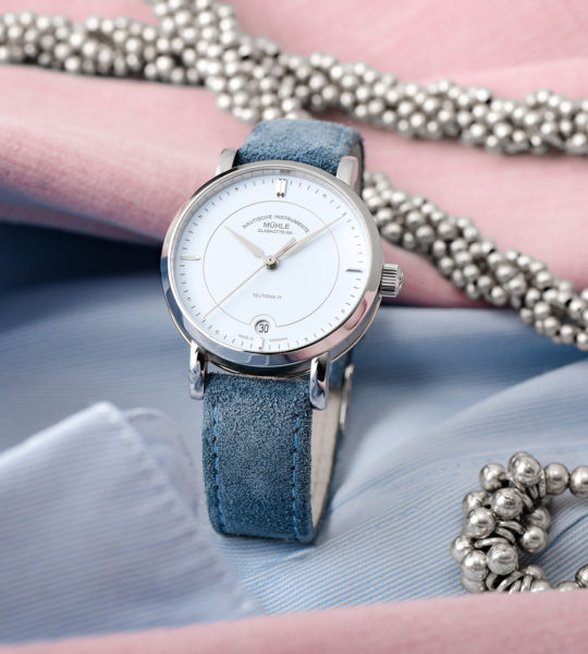 Armbanduhr Teutonia IV Lady mit pastellblauem Zifferblatt am blauen Veloursband steht auf hellblauem und rosafarbenem Stoff mit silbernem Schmuck.