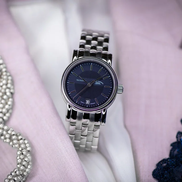 Armbanduhr Teutonia II Medium mit blauem Zifferblatt am Metallband steht auf rosa und weißen Stoffen.