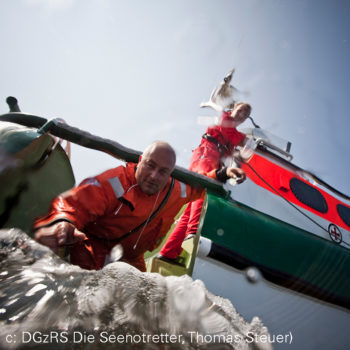 Rettungseinsatz Seenotretter: Ein Mann mit roter Rettungskleidung in einem kleinen Boot greift nach vorn, im Hintergrund ein Seenotrettungsboot.