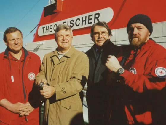 Vier Männer, darunter Hans-Jürgen Mühle (2.v.l.) bei der Übergabe des ersten S.A.R. Rescue-Timer. Im Hintergrund der Seenotrettunskreuzer "Theo Fischer".