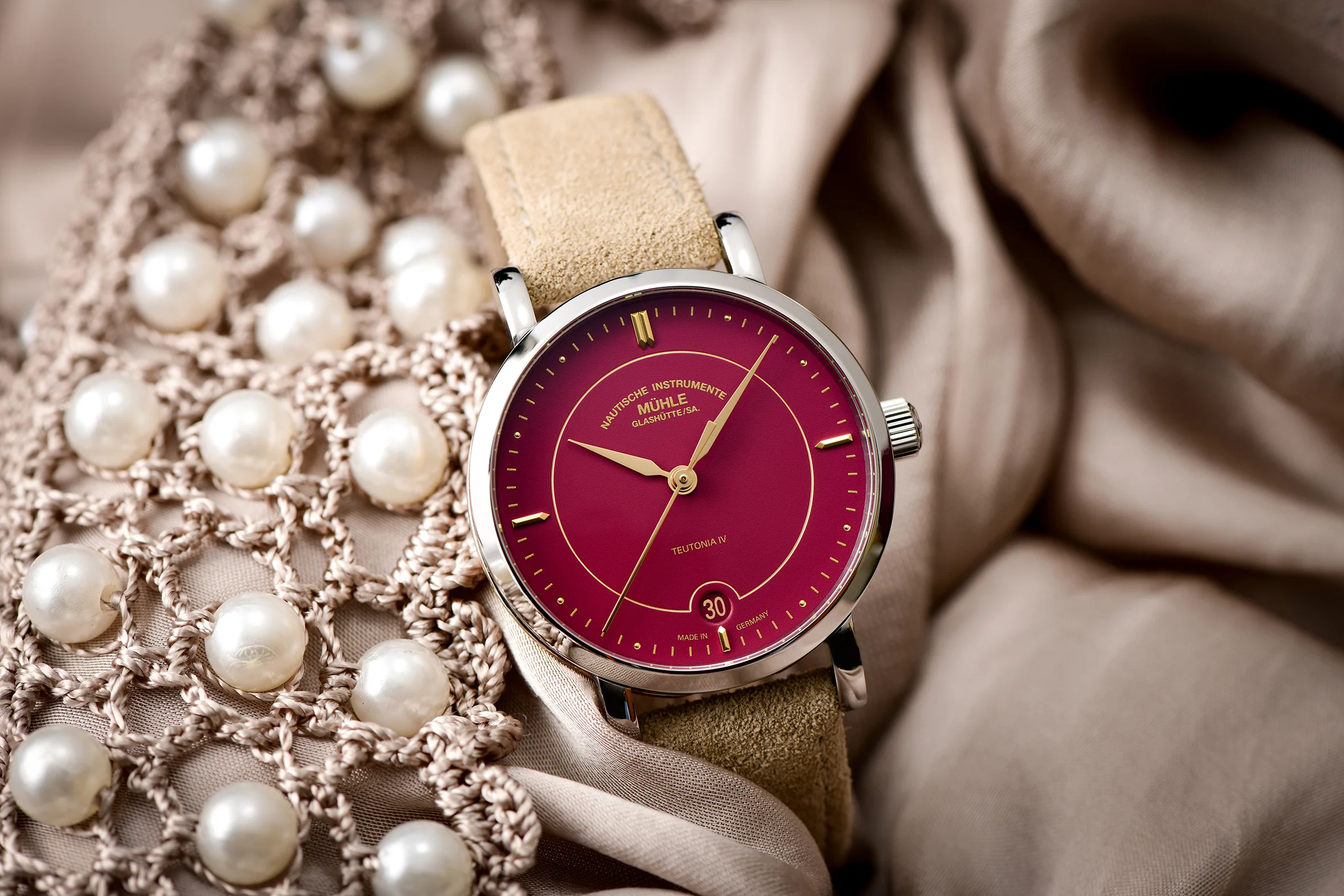 Armbanduhr Teutonia IV Lady mit himbeerrotem Zifferblatt am beigen Veloursband steht zwischen beigem Stoff und Perlenschmuck.