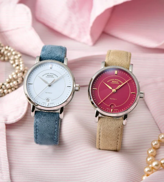Zwei Armbanduhren (Teutonia IV Lady mit pastellblauem Zifferblatt am blauen Veloursband und mit himbeerrotem Zifferblatt am beigen Veloursband) stehen nebeneinander auf einer rosafarbenen Damenbluse.