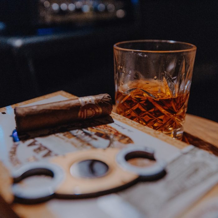 Eine Zigarrenschachten, darauf eine Zigarre und ein Metallteil, liegen auf einem Tisch. Dahinter steht ein Cognac im Kristallglas.