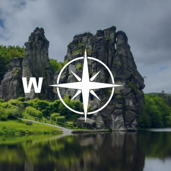 Kompassrose, das auf den Buchstaben "W" zeigt. Im Hintergrund ein zerklüfteter Fels an einem Fluss.