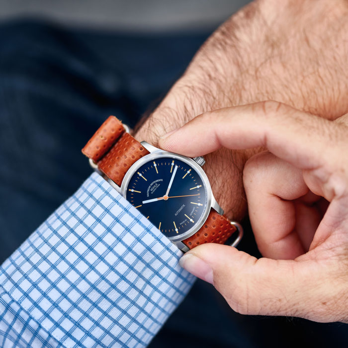 Wristshot Panova blau: Armbanduhr mit blauem Zifferblatt und silbernem Gehäuse (Panova blau) am braunen Lederband an einem Handgelenk vor einem blau-weiß karierten Hemdsärmel.
