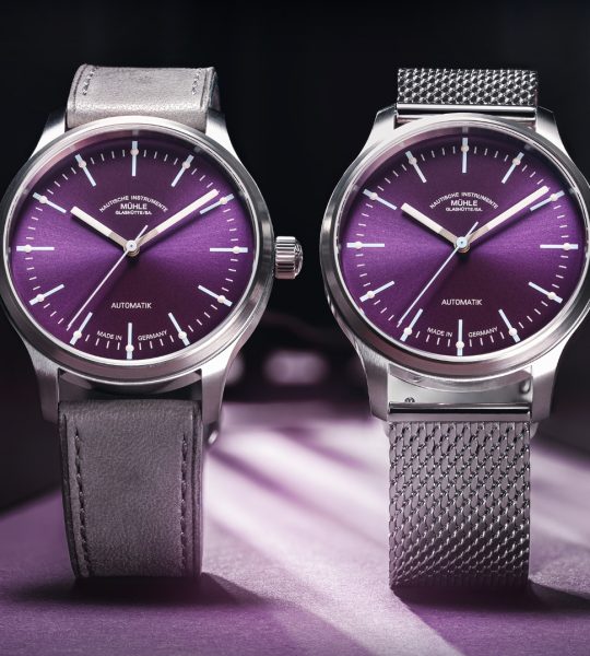Zwei Armbanduhren (Panova Violett am grauen Lederband und am Milanaiseband) stehen nebeneinander auf einer violetten Oberfläche.