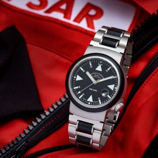 S.A.R. Rescue-Timer mit schwarzem Zifferblatt und Metall-/Kautschukband steht auf einer roten Jacke mit SAR-Aufnäher.