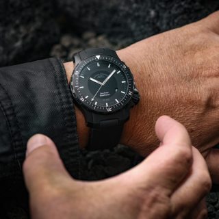 Armbanduhr Sea-Timer BlackMotion am schwarzen Kautschukband wird am Handgelenk getragen. Eine weitere Hand greift nach der Uhr.