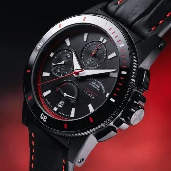 Leicht schräge Ansicht des Sportivo Power Chronograph First Edition am schwarz-roten Lederband vor rotem Hintergrund