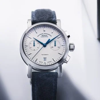 Teutonia IV Chronograph mit weißem Zifferblatt im silbernen Gehäuse am blauen Velourslederband steht auf einem weißen Untergrund.