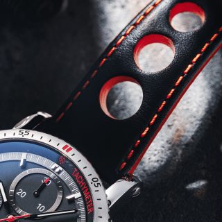 Makroaufnahme eines schwarz-roten Lederbandes an einer Armbanduhr mit schwarzem Zifferblatt und silbernem Gehäuse.