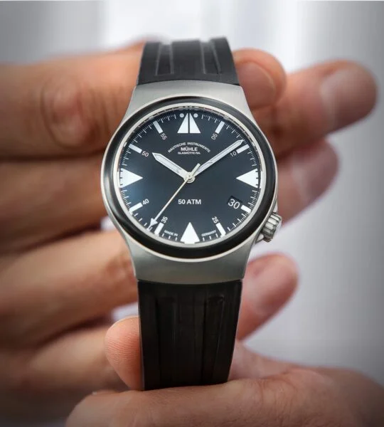 S.A.R. Mission-Timer – eine Armbanduhr mit schwarzem Zifferblatt, weißen Indizes, mattsilbernem Gehäuse und schwarzem Kautschukband – wird von zwei Händen gehalten.