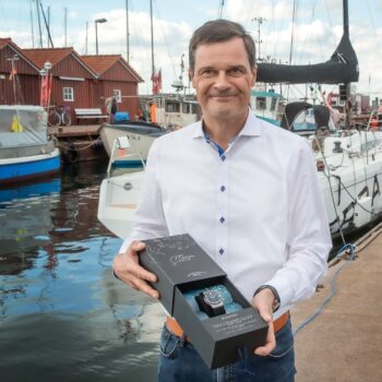 Thilo Mühle hält einen S.A.R. Mission-Timer in einer schwarzen Box in der Hand. Im Hintergrund ein Hafen mit Schiffen und braunen Häusern.